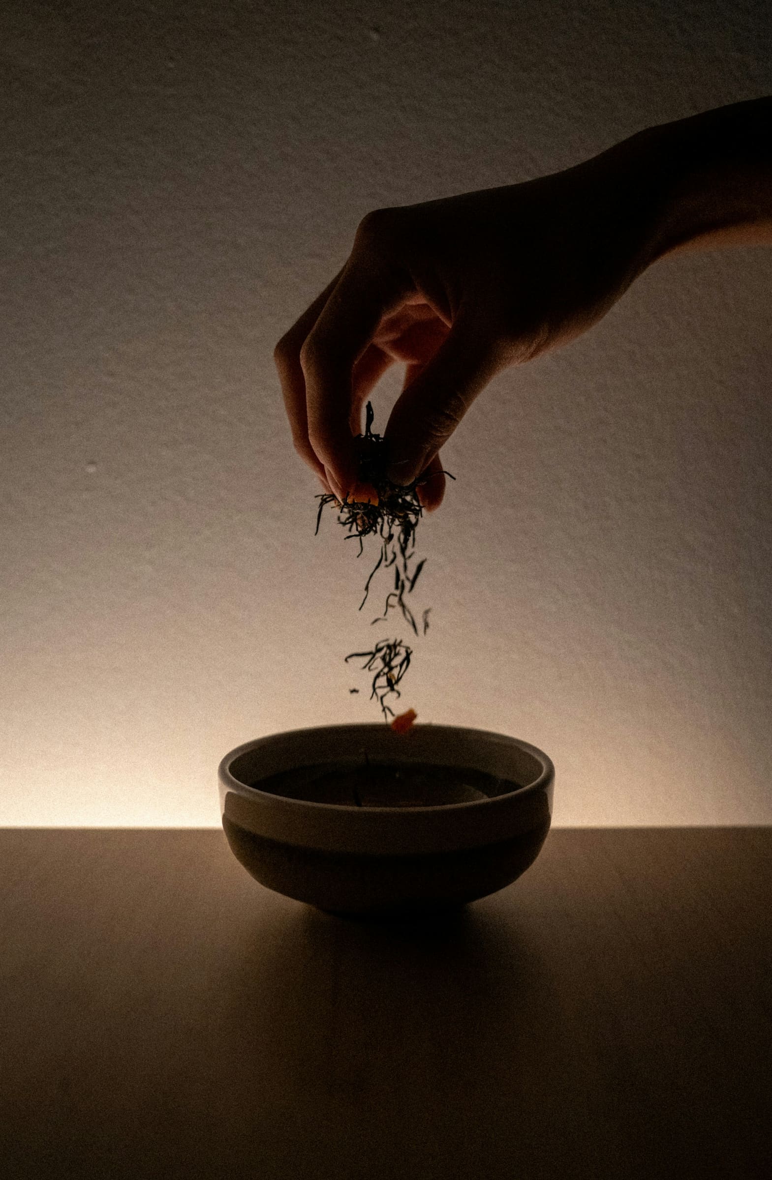 A hand scattering loose tea leaves into a dark bowl